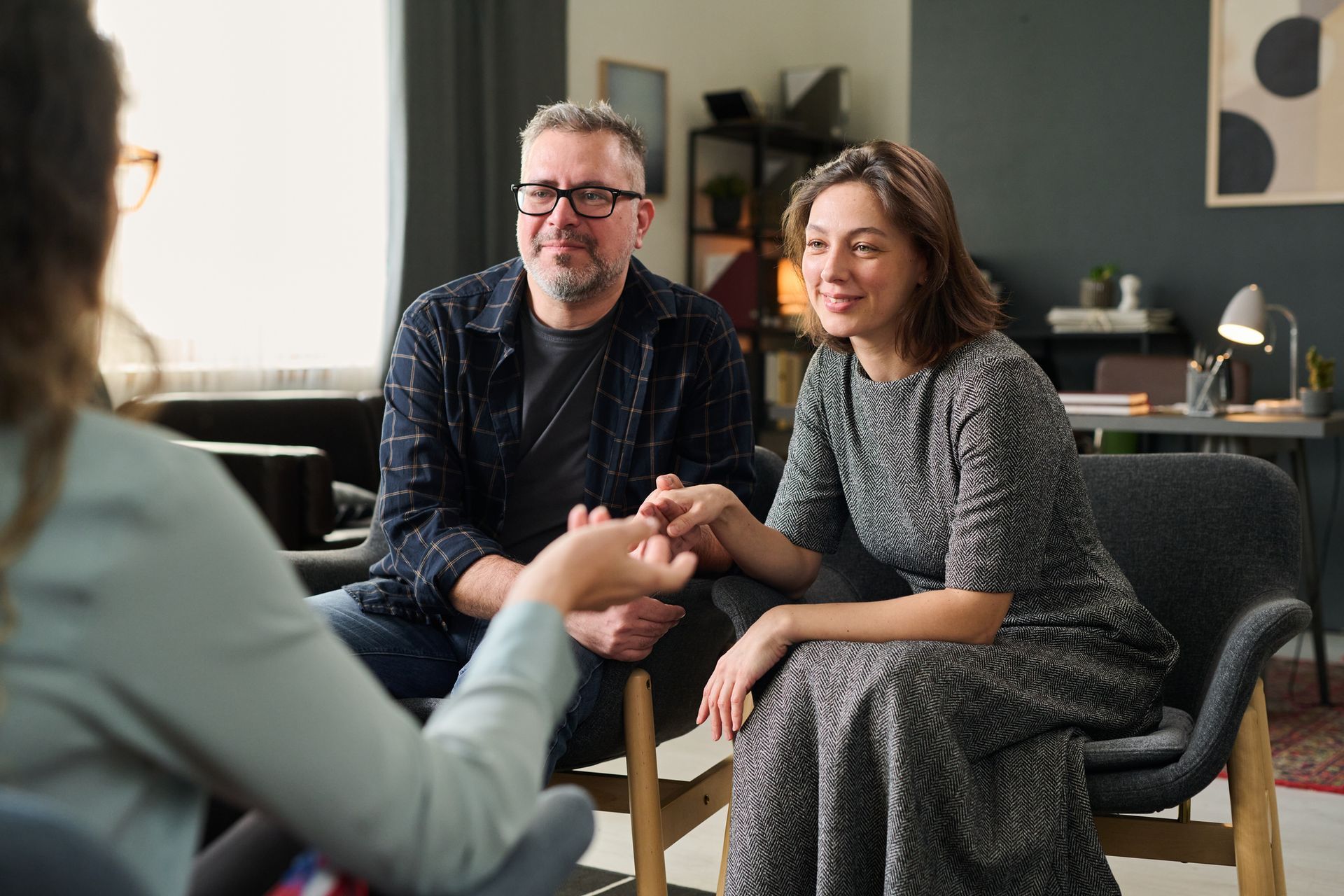 Couple in therapy session, holding hands, with counselor in a cozy office setting.