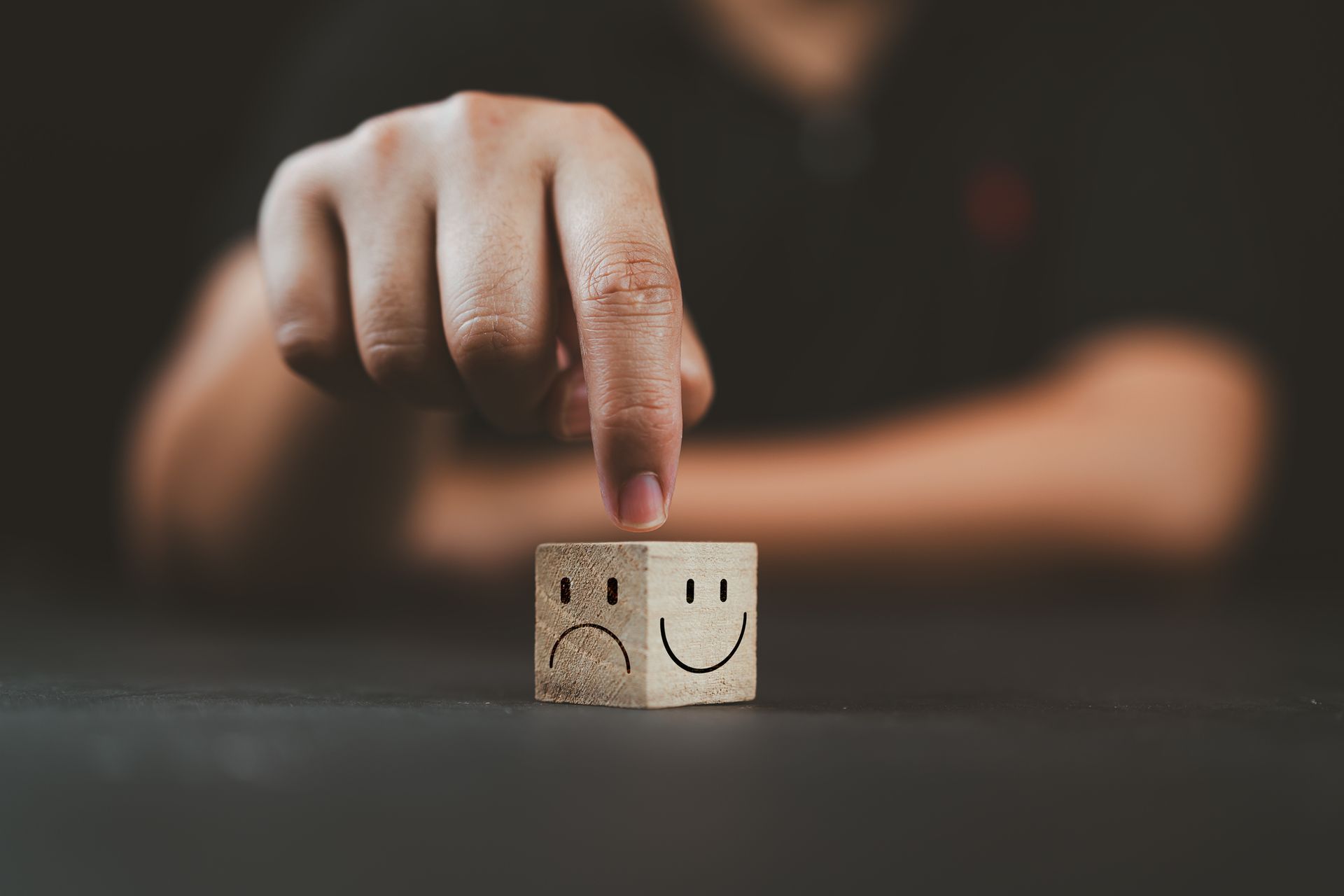 A person's finger points at a wooden block showing a sad face on one side and a happy face on the other.