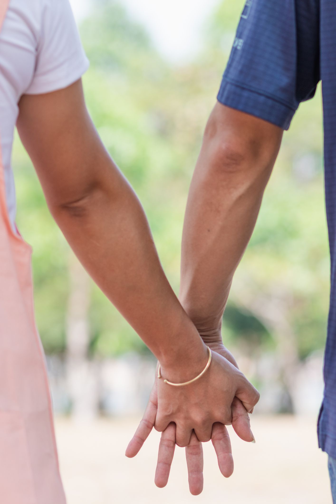 Two people holding hands, outdoors. One wears pink, the other blue. Sunlight, blurred background.