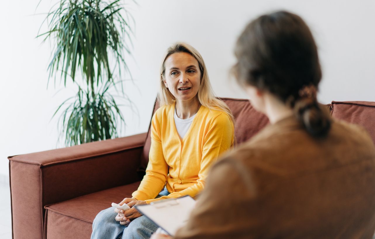 Woman in yellow cardigan on a red couch speaking to person holding clipboard; indoor setting with a plant.