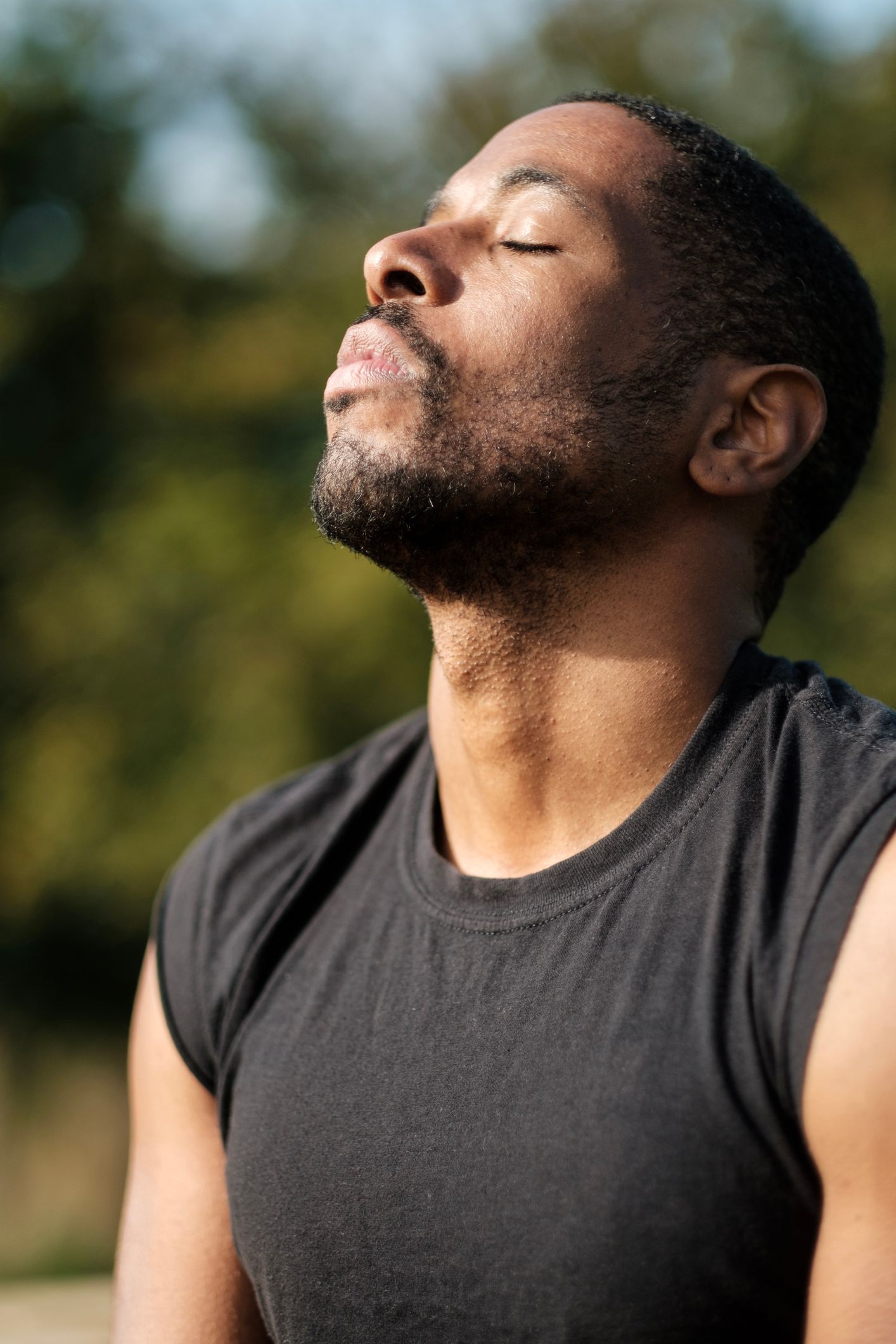 Man with closed eyes, sweat on face, looking up. Wearing black sleeveless shirt, outdoors with blurred green background.