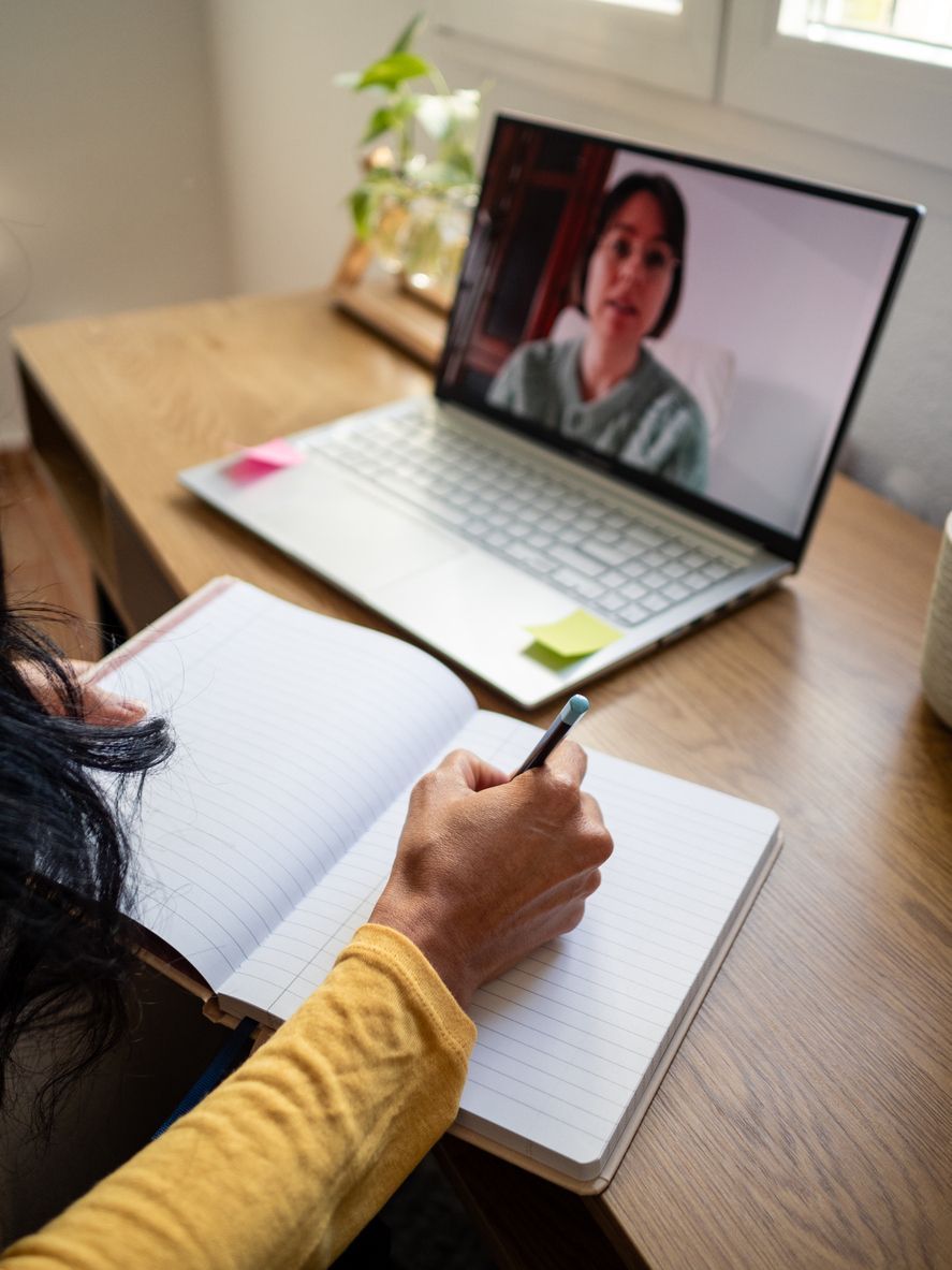 Person taking notes during a video call on a laptop. Notebook open, pen in hand, woman on screen.