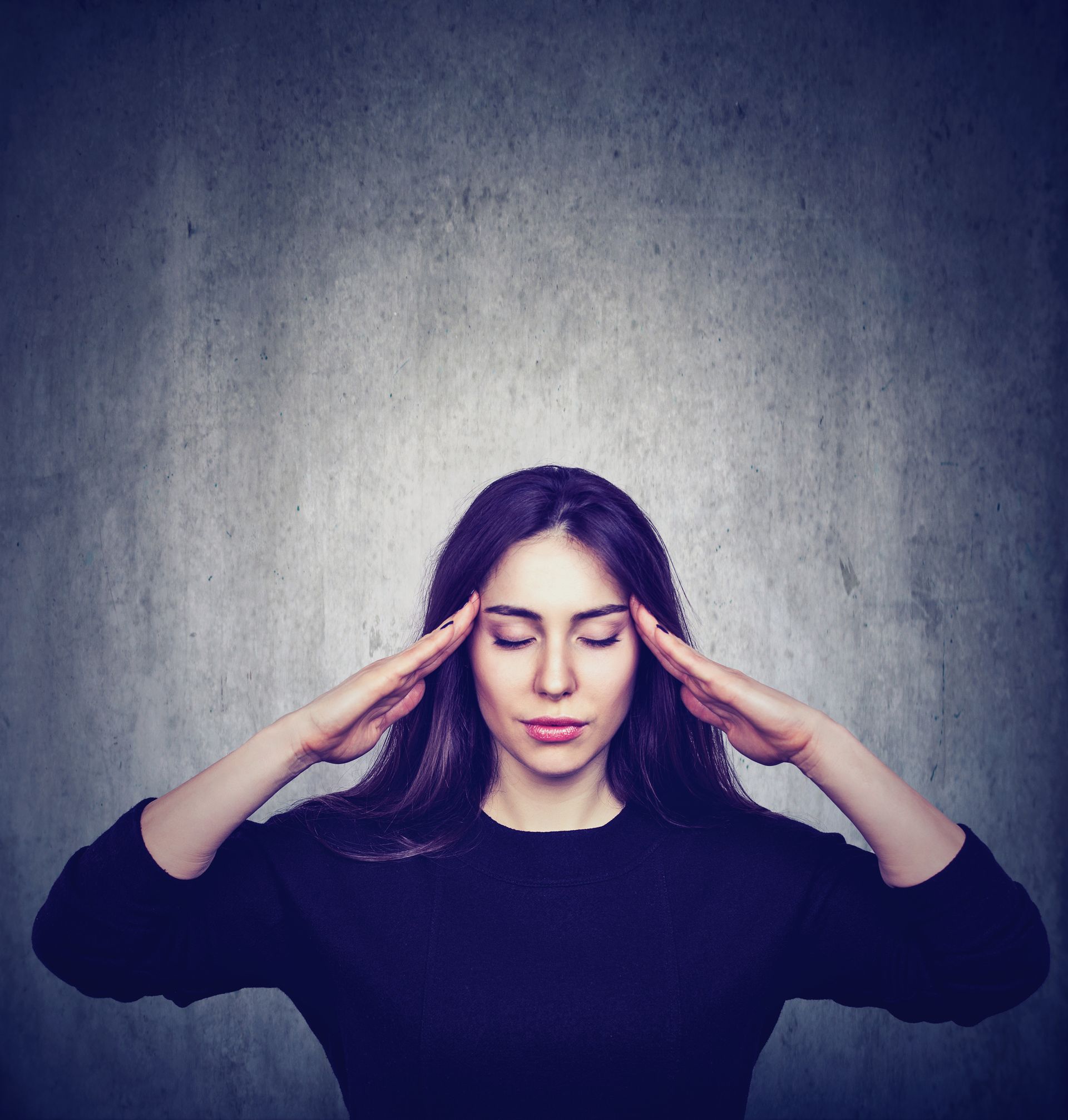 Woman with dark hair, eyes closed, hands on temples, looking stressed. Against gray background.