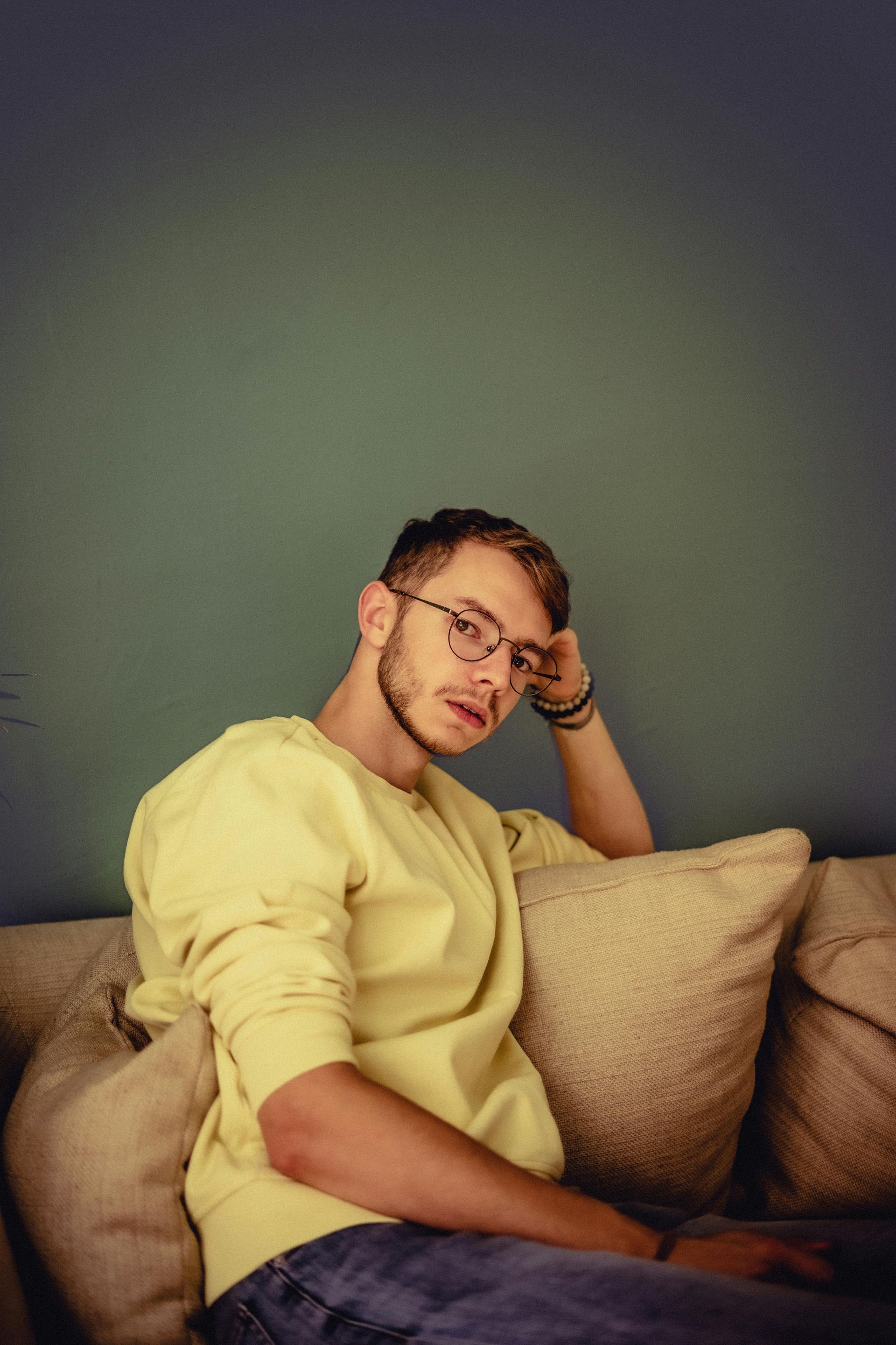 Man with glasses and yellow shirt lounging on a couch, resting his head.
