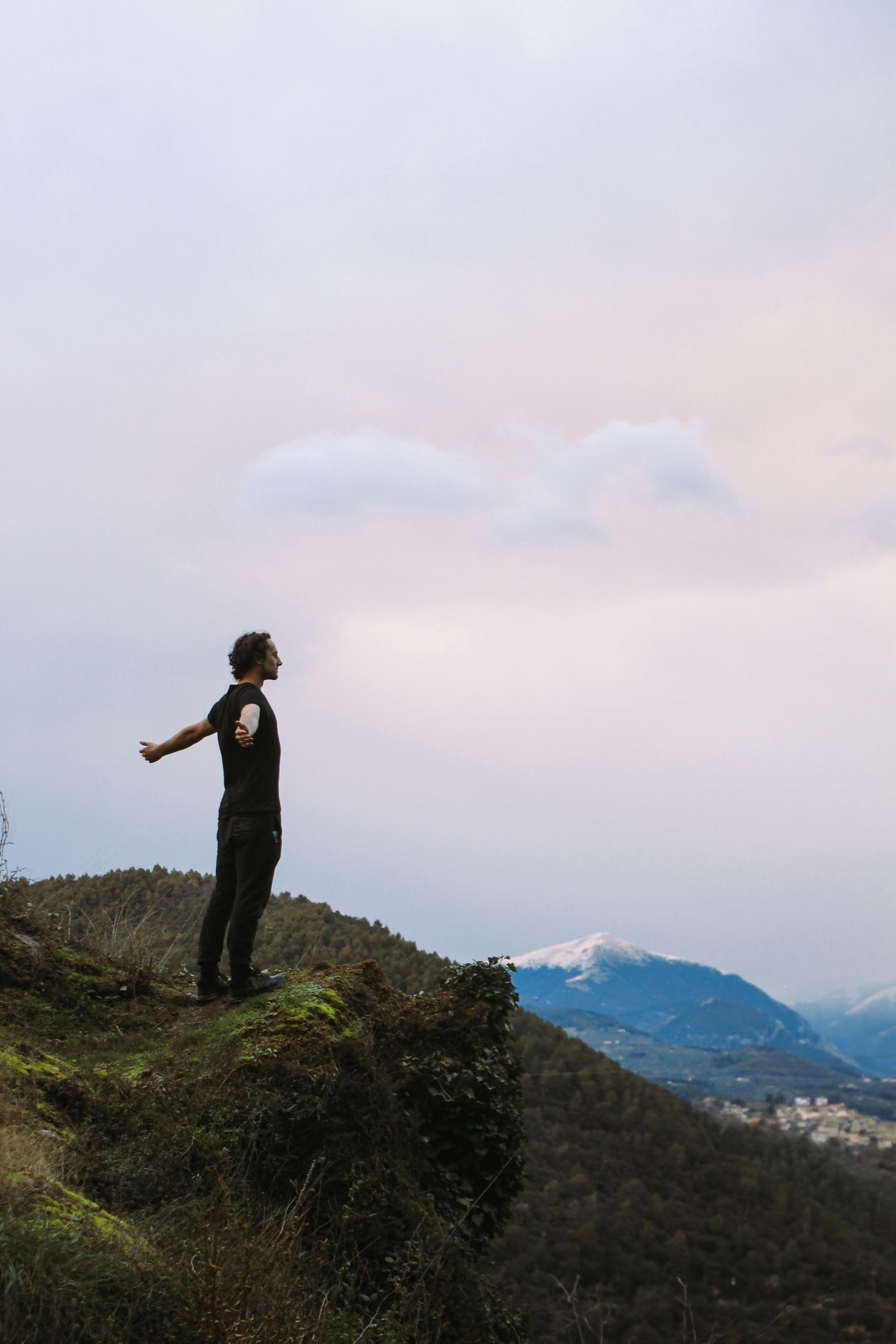 Man with arms outstretched on a cliff, overlooking mountains under a cloudy sky.