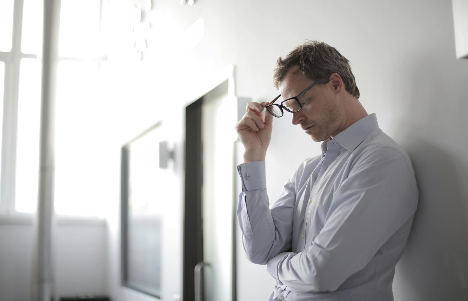 Man in a light blue shirt leans against a wall, holding glasses, looking down, and appearing contemplative.
