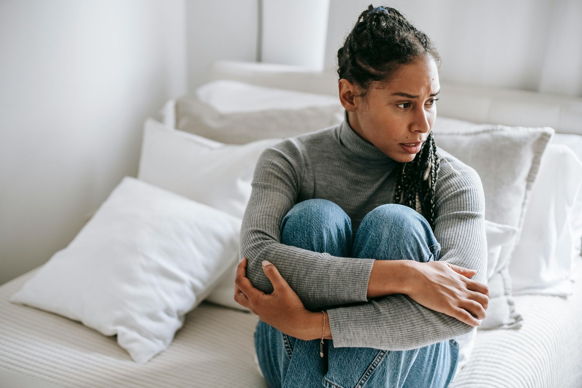 Woman with braids sits on bed, hugging knees, looking concerned.