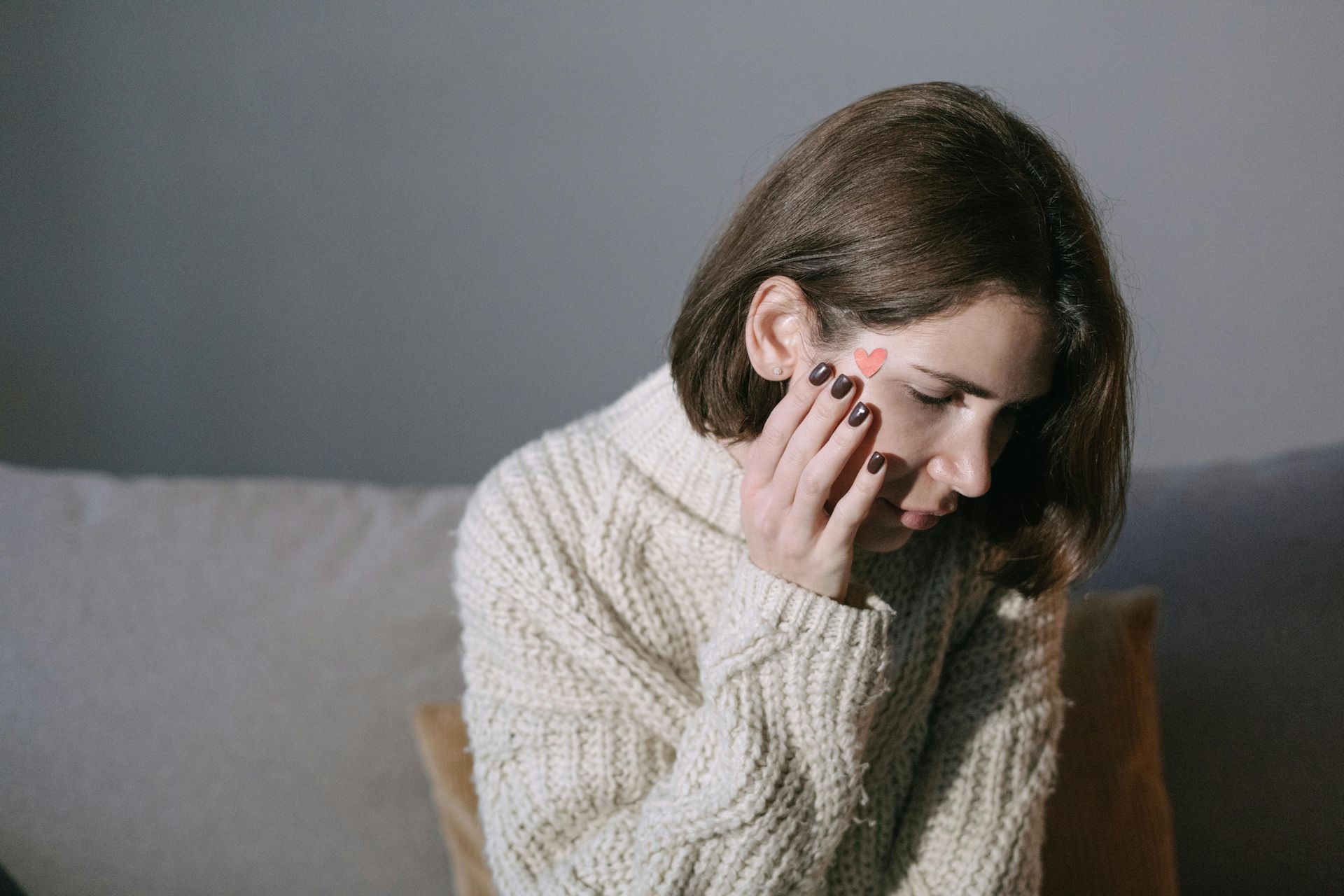 Woman in cream sweater touches face, a red heart-shaped bandage on her cheek.