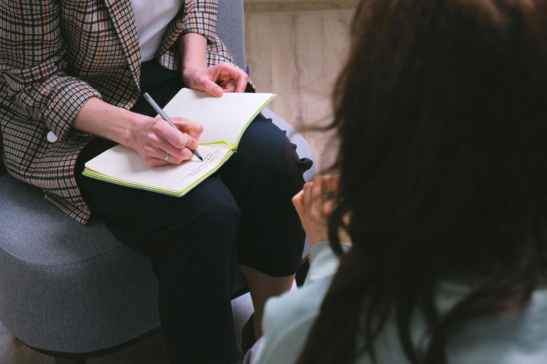 Person writing in notepad while facing another person seated in a chair.