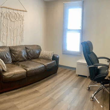 Brown leather sofa, macrame wall hanging, office chair, and window in a room.