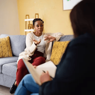 Woman talking, gesturing on a sofa, facing someone holding a notepad. Yellow wall, neutral setting.