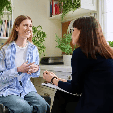 Woman in a striped shirt talks to a person holding a clipboard in an office setting with plants and bookshelves.