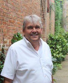 A man in a white shirt is standing in front of a brick wall.