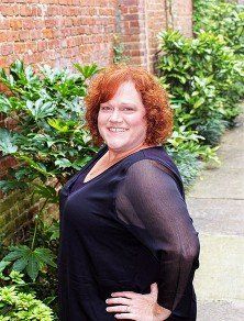 A woman with red hair is standing in front of a brick wall with her hands on her hips.