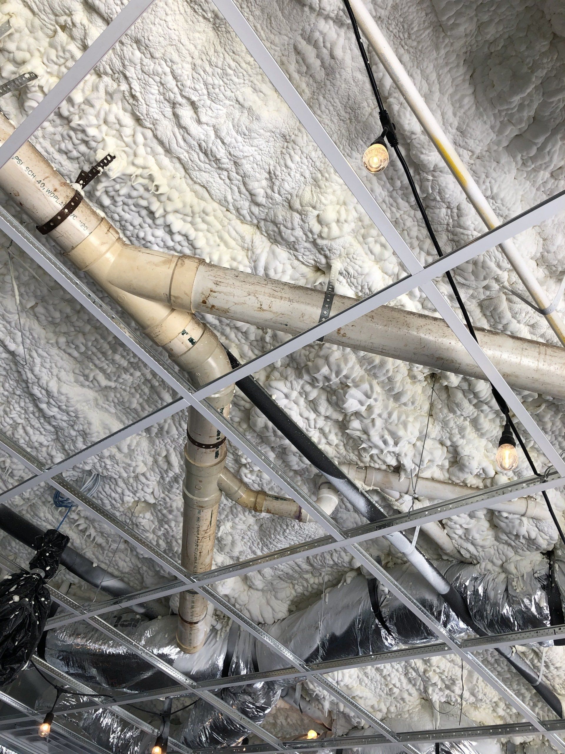 spray foam above a bunch of pipes are hanging from the ceiling of a building .
