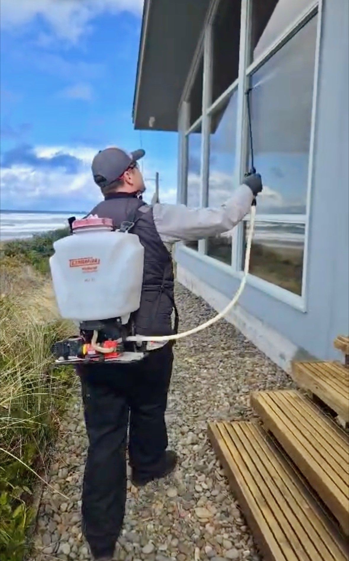 Person spraying window with backpack sprayer by a beach house.
