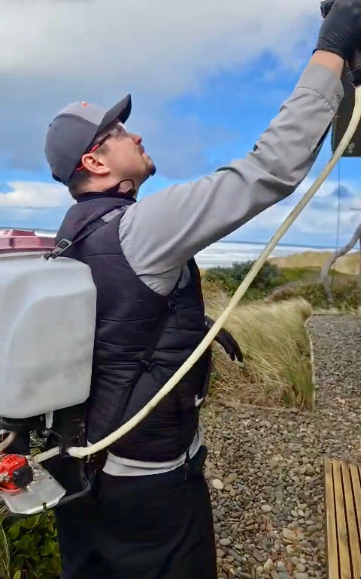 Man in cap sprays with backpack sprayer, outdoors near beach and buildings.