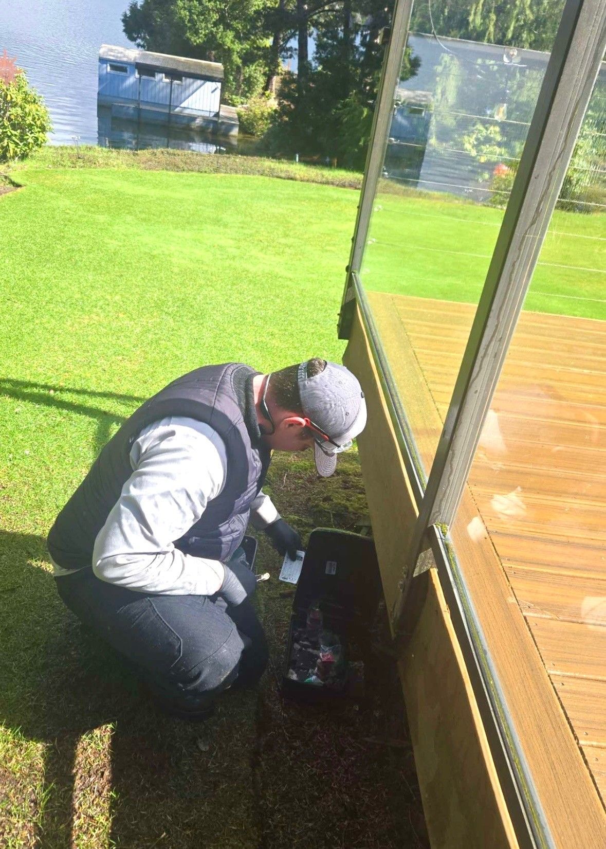 Man kneeling, inspecting something near a deck with glass panels, on grassy lawn by lake.