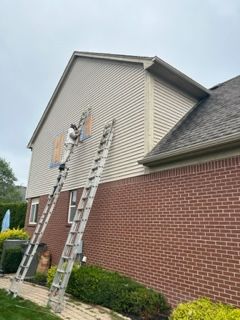 A man is standing on a ladder painting the side of a brick house.