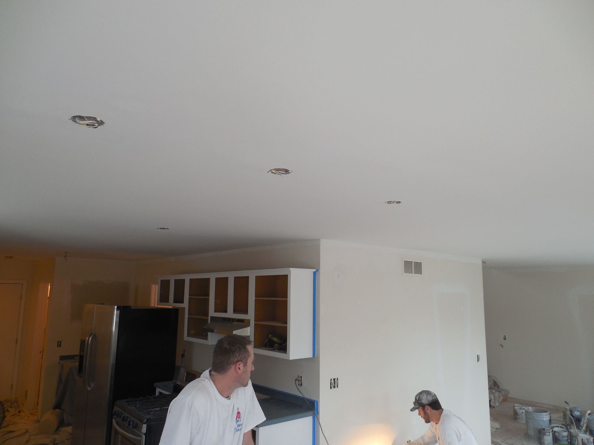 Two men are painting a kitchen with white cabinets.