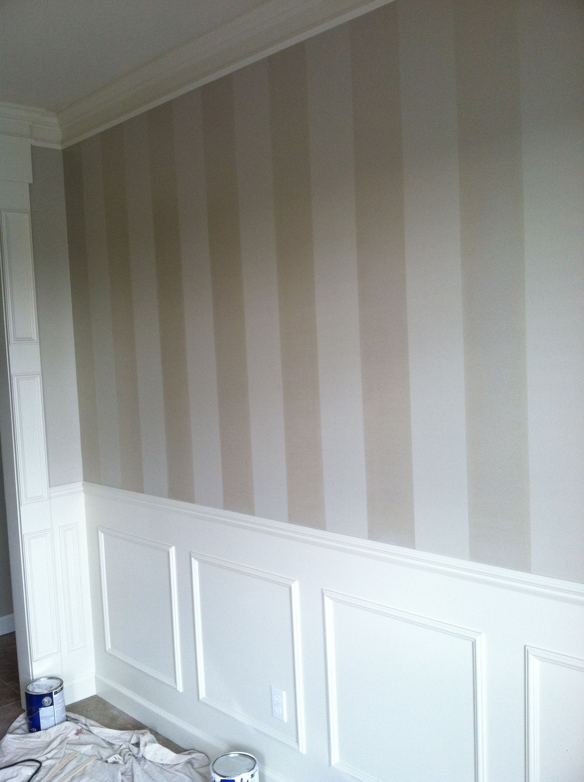 A wall with a striped wallpaper and white trim is being painted.
