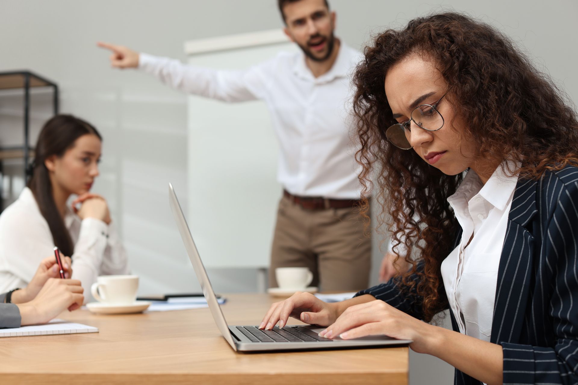 A professional team in an office setting; one person types at a laptop while another points toward a colleague in the back.