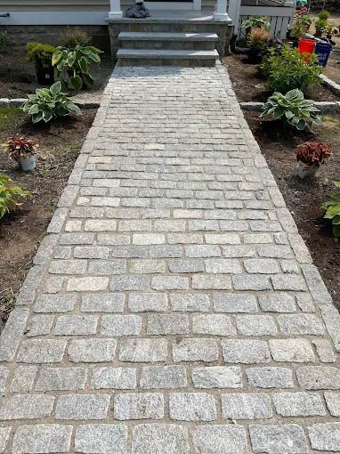 Granite stone walkway leading to a house entrance, lined with planted flower beds.