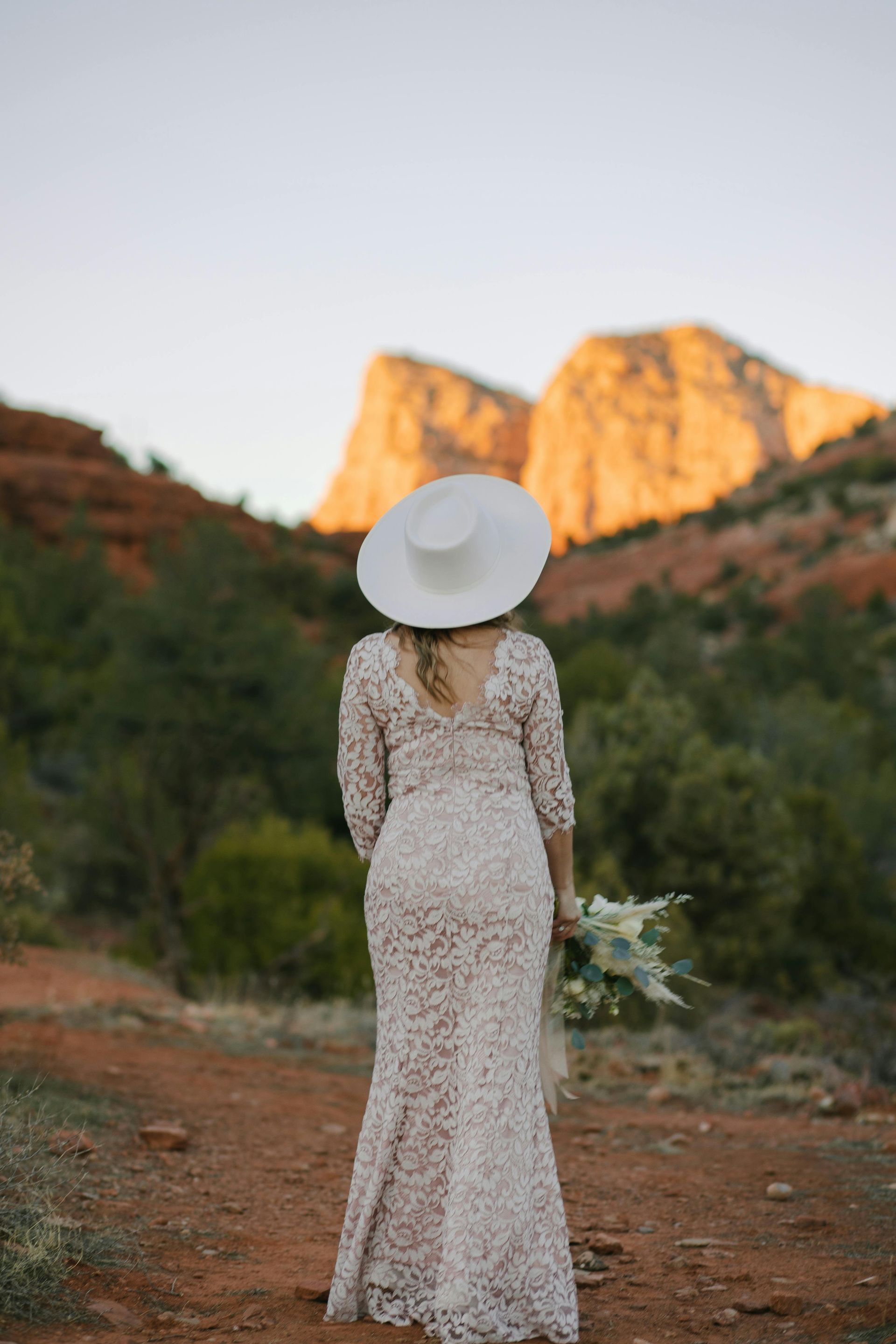 A woman in a wedding dress and hat is standing in front of a mountain.