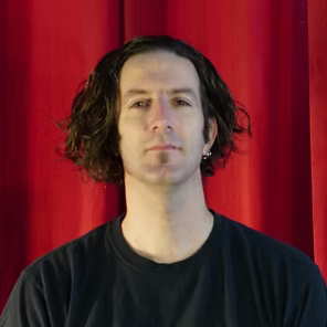 Person with shoulder-length dark hair and black shirt in front of a red curtain