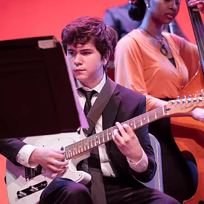 Musician in a suit playing electric guitar on stage with a red-lit background