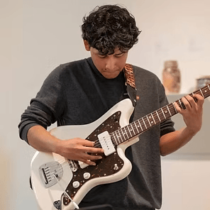 Person playing a white electric guitar indoors, looking down while strumming.