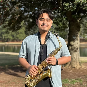 Man holding a saxophone outdoors in a park by a lake, wearing a light blue shirt.