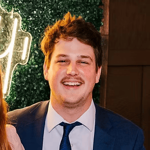 Smiling man in a blue suit and tie posing against a leafy backdrop with warm lights