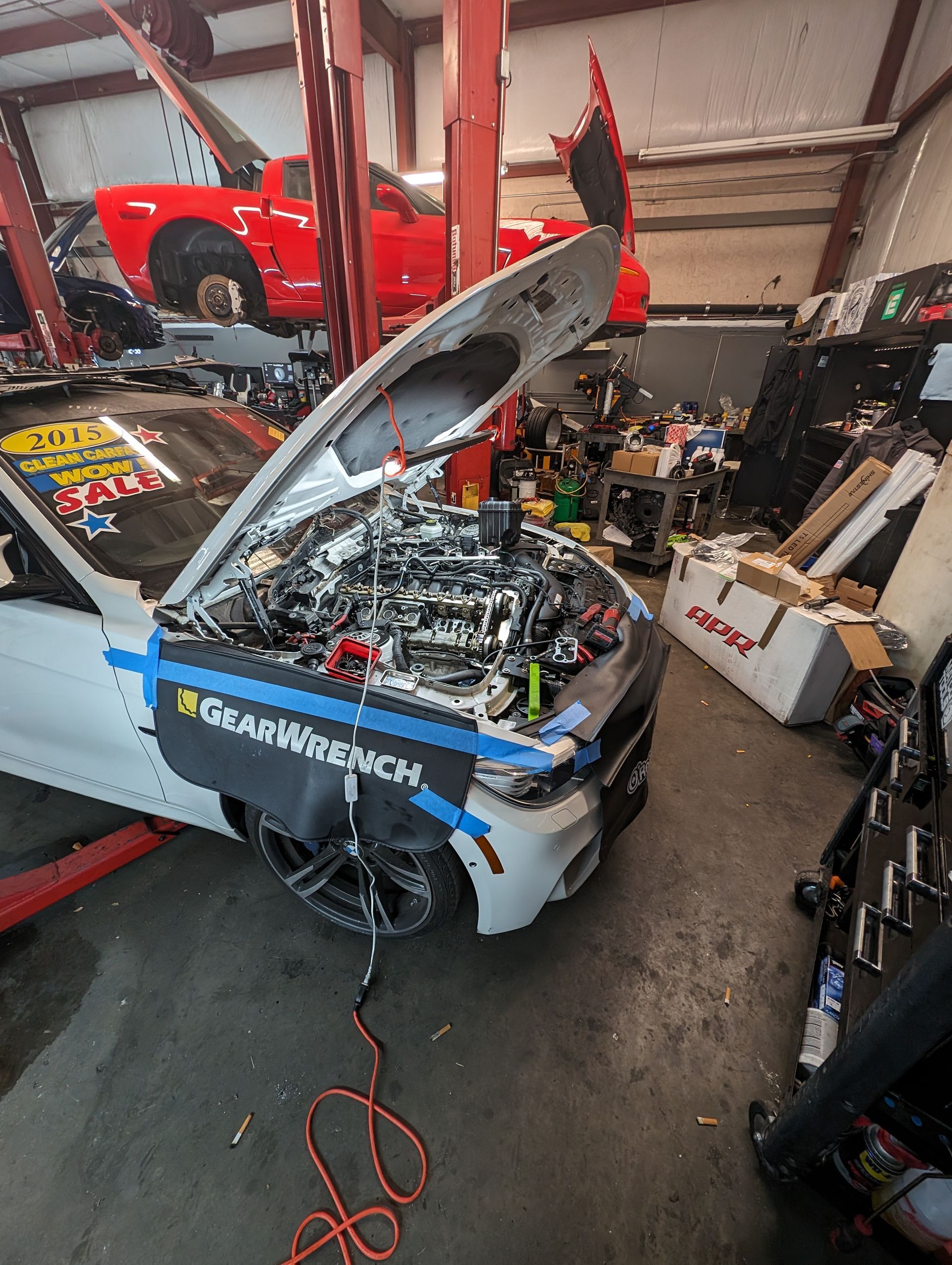 A white car with open hood, being worked on in a garage. Red car on lift in background. | Platinum Motors Martinez