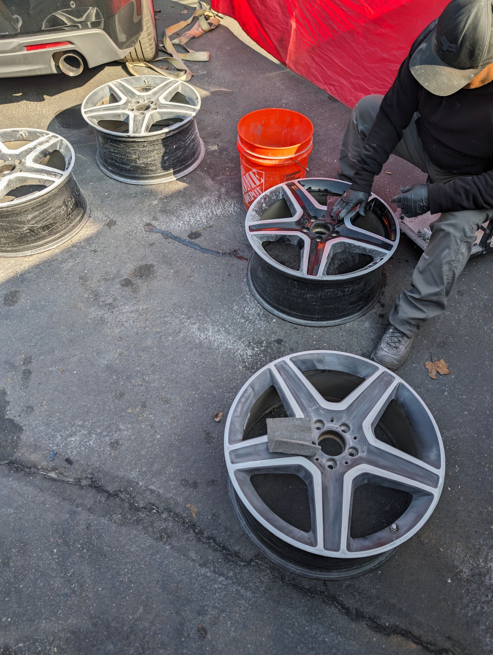 Person sanding a car wheel with several others nearby; orange bucket on asphalt.  | Platinum Motors Martinez