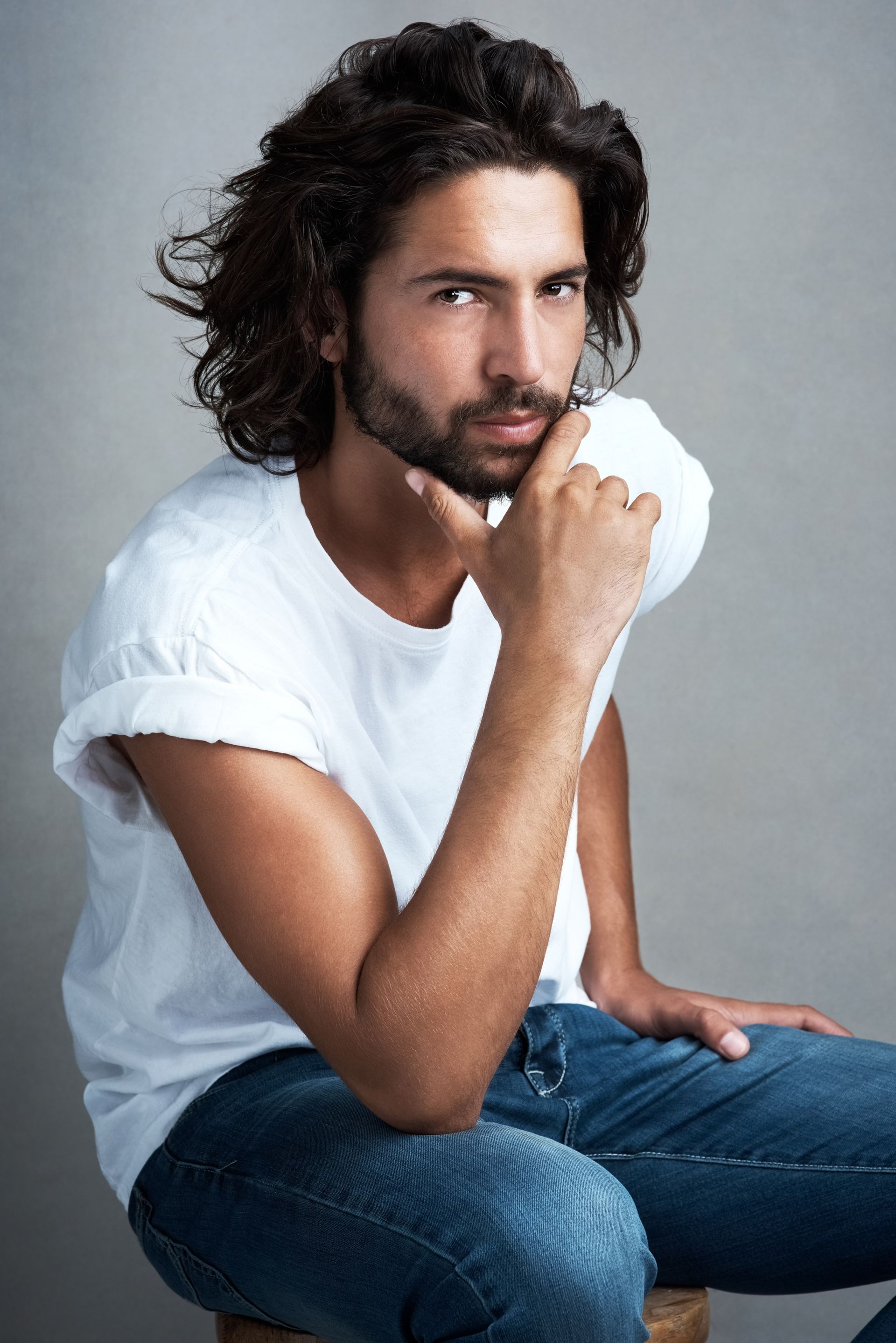 A man with long hair and a beard is sitting on a stool with his hand on his chin.