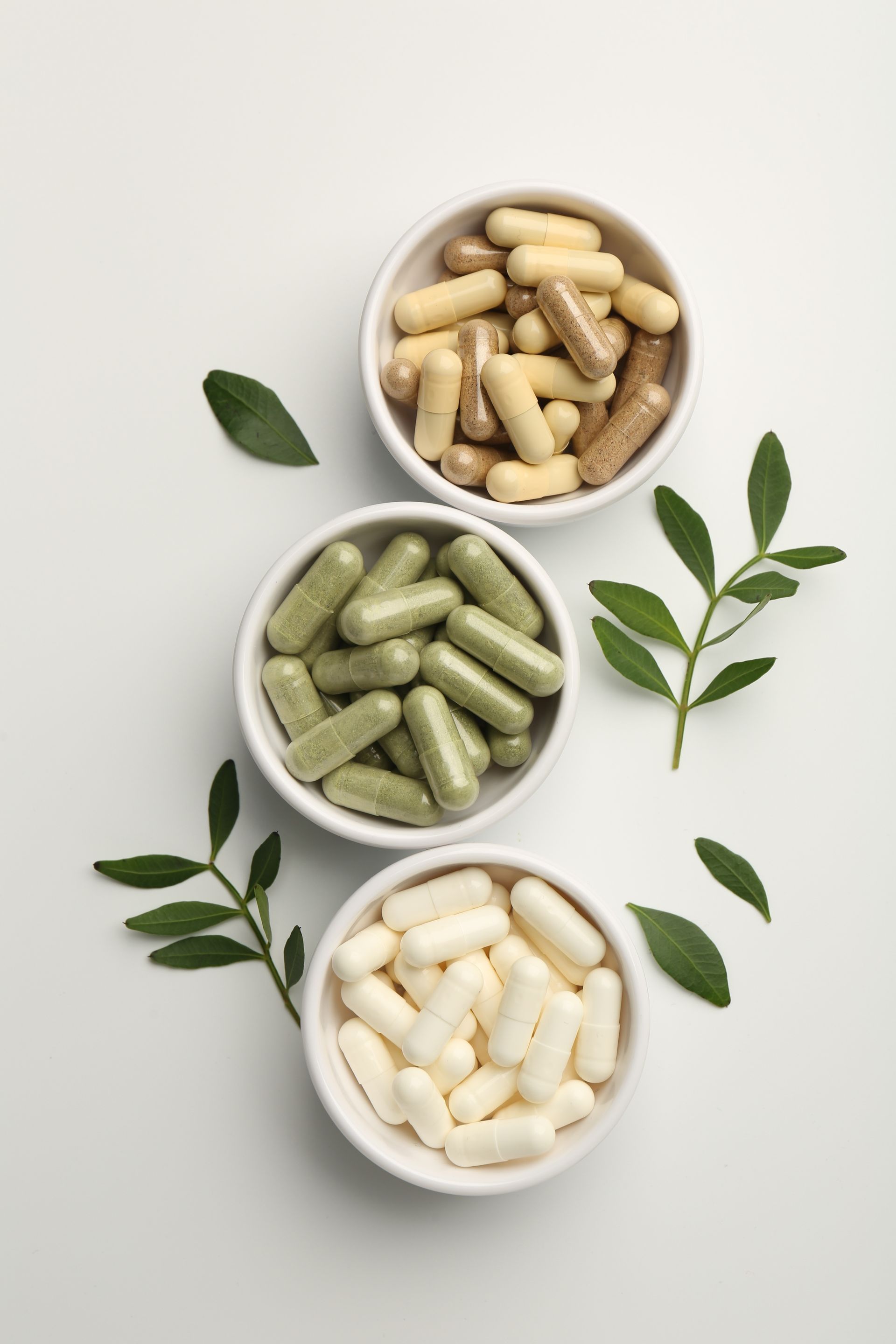 Three bowls filled with different types of pills and leaves on a white background.