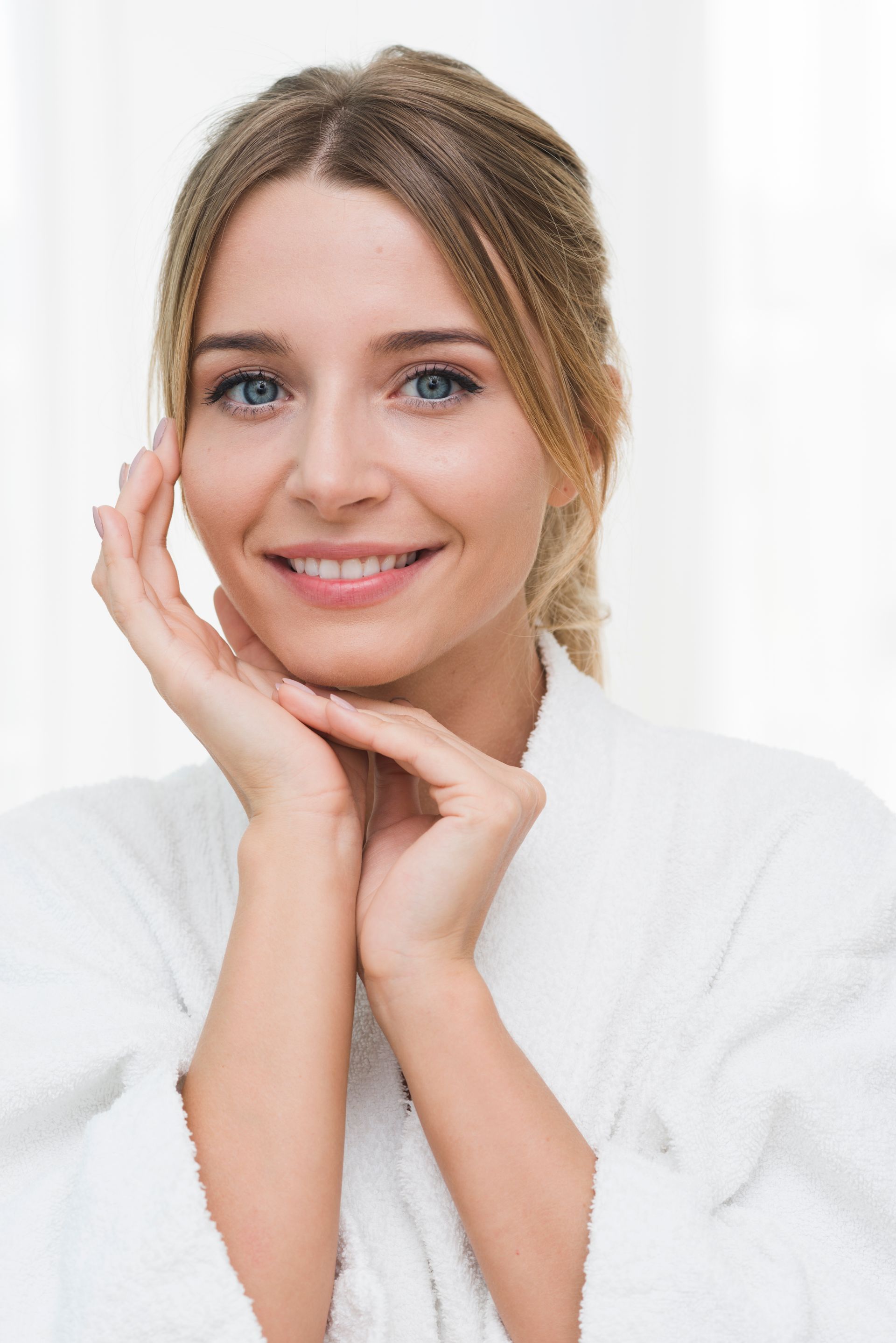 A woman in a bathrobe is smiling and touching her face.