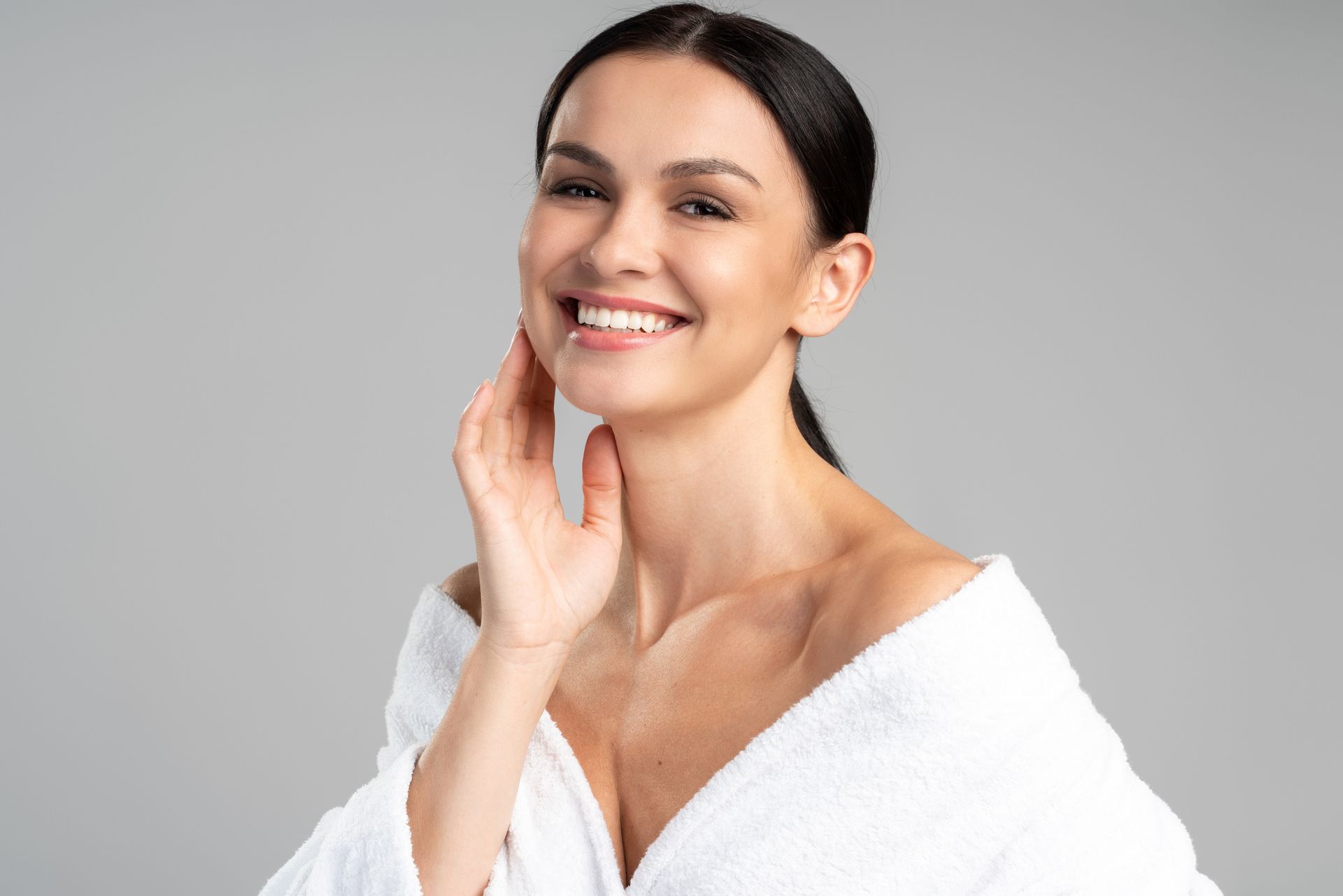 A woman in a white towel is smiling and touching her face.