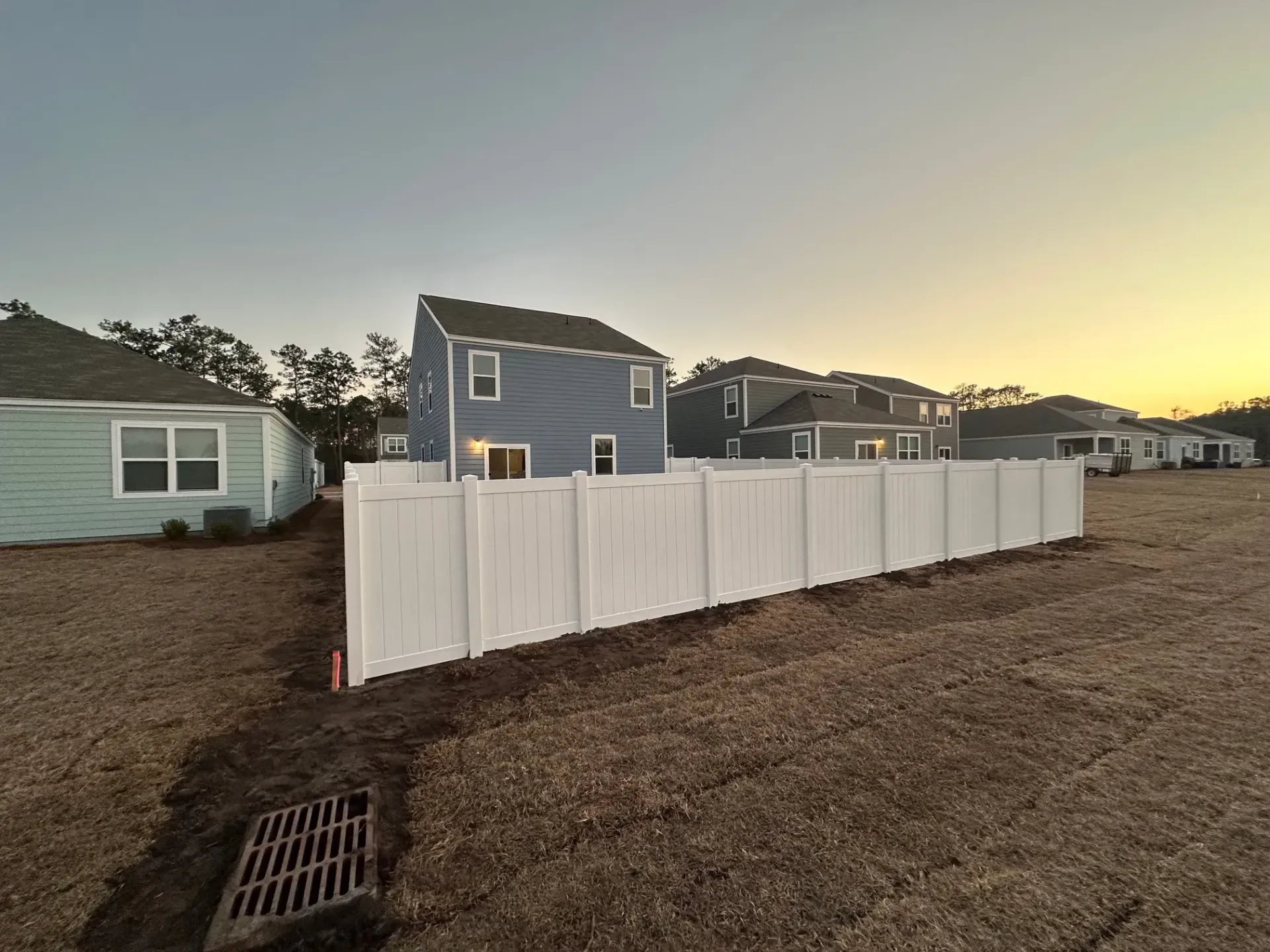 White fence in front of blue house, other houses in the background at dusk.
