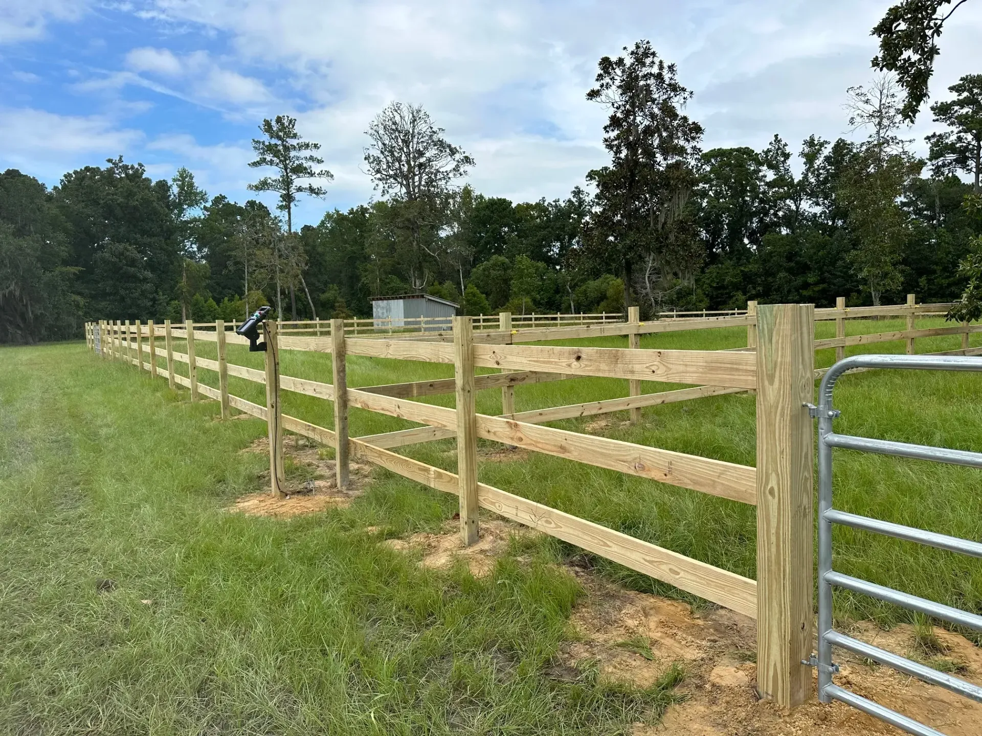 Wooden fence in a grassy field, with a metal gate open. Trees and a small building are in the background.