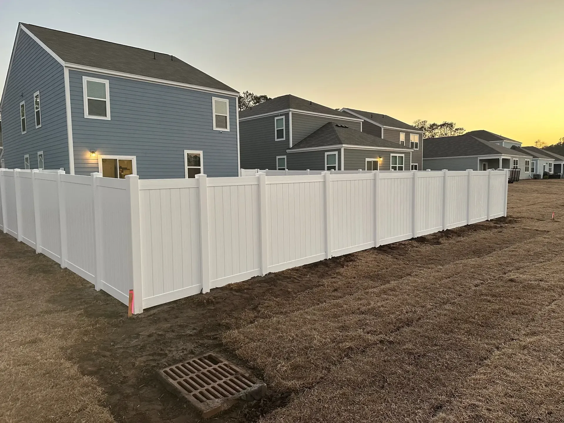 White vinyl fence around a blue house and adjacent houses in an open lot.