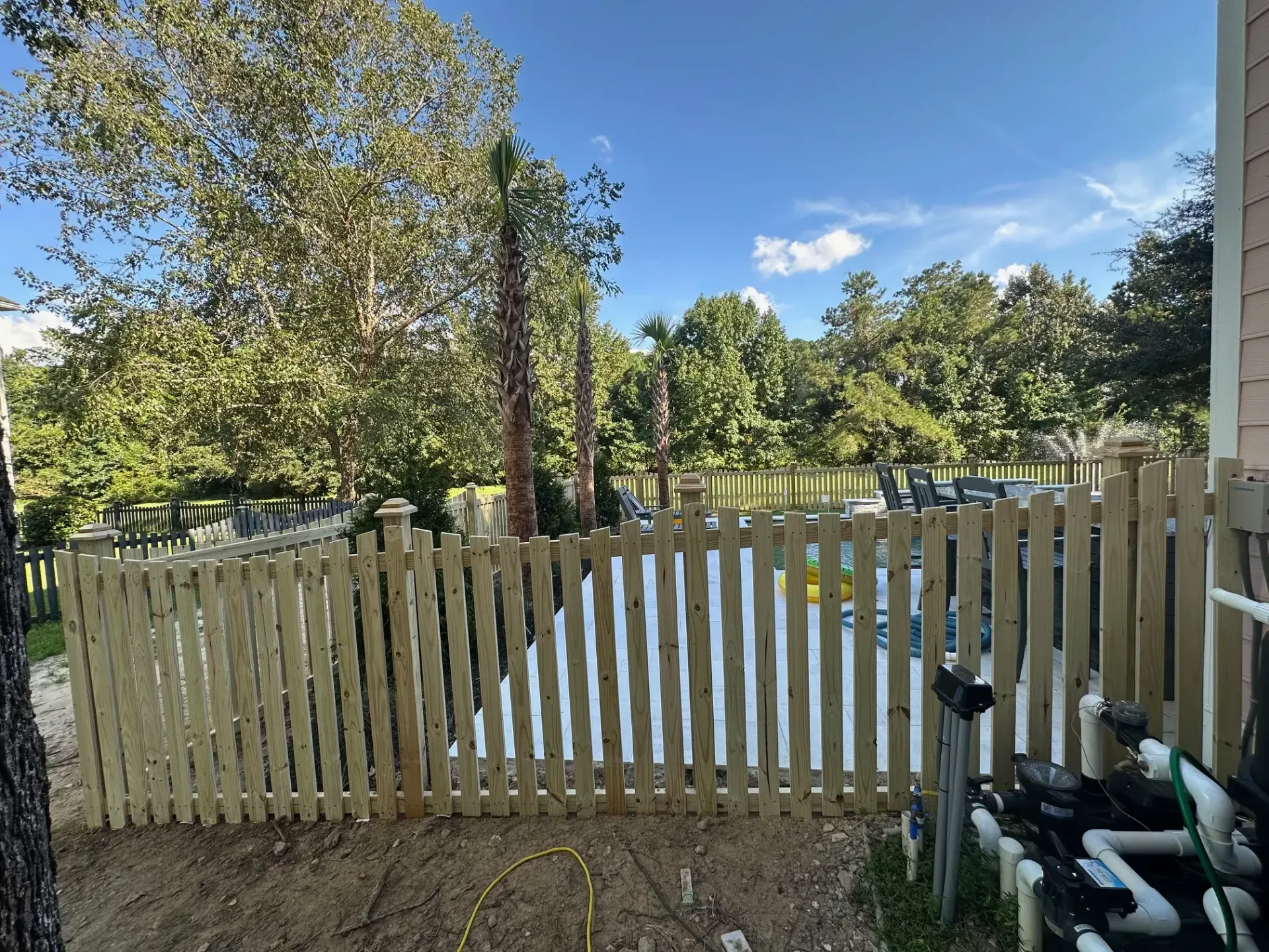 Wooden fence surrounding a swimming pool in a backyard with trees and blue sky.