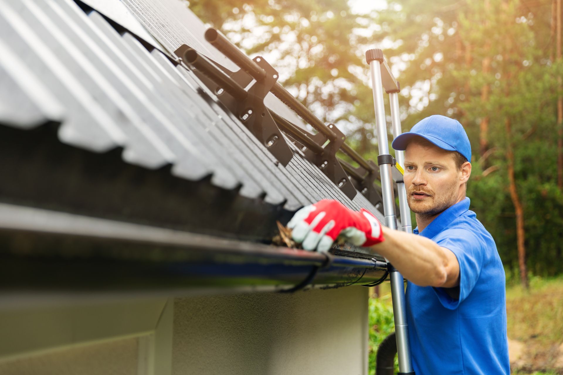 Worker cleaning gutters on a house roof with a long tool, wearing blue uniform and red gloves