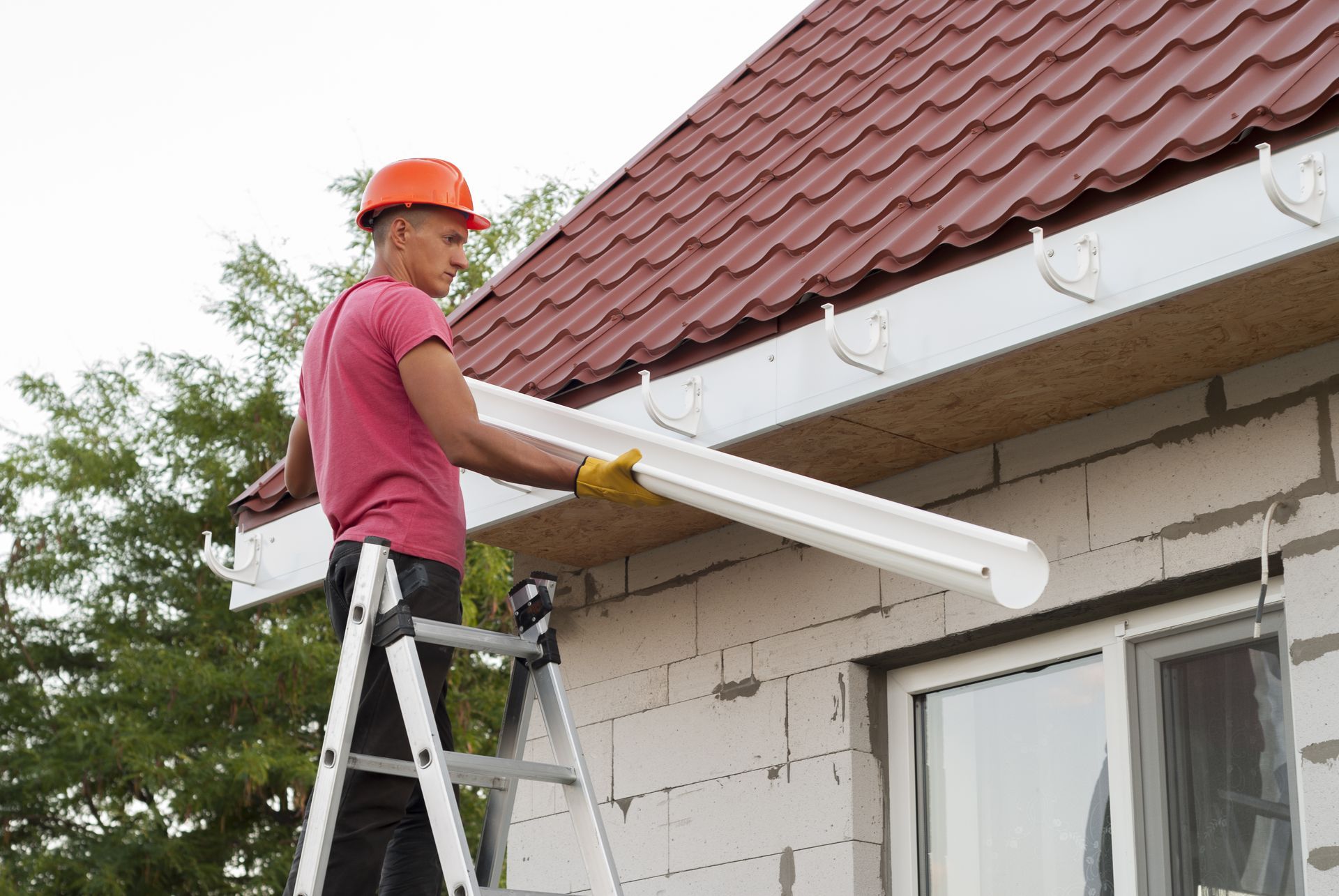 Worker on a ladder installing a white gutter on a house roof edge