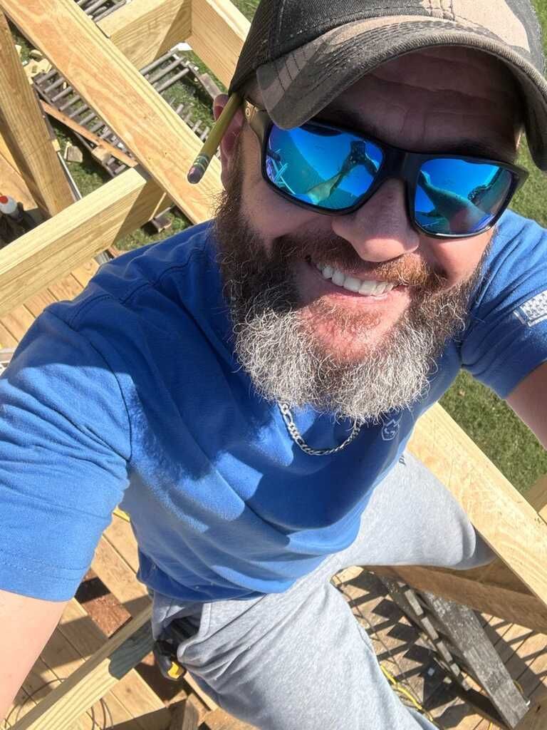 Man wearing sunglasses and a hat smiles, positioned on a wooden structure, possibly construction or carpentry.