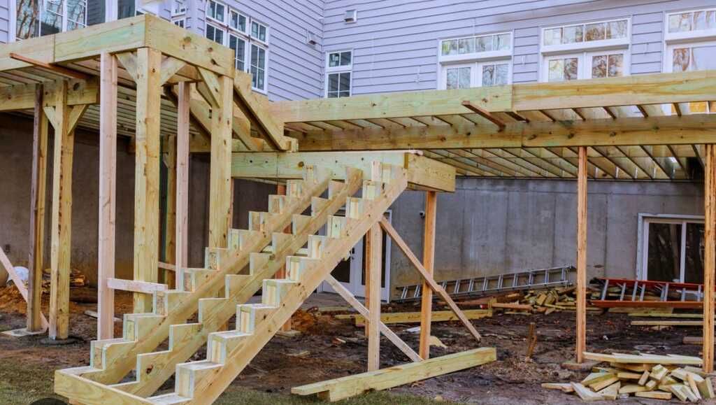 Wooden deck construction in progress next to a house with unfinished siding. Staircase visible.