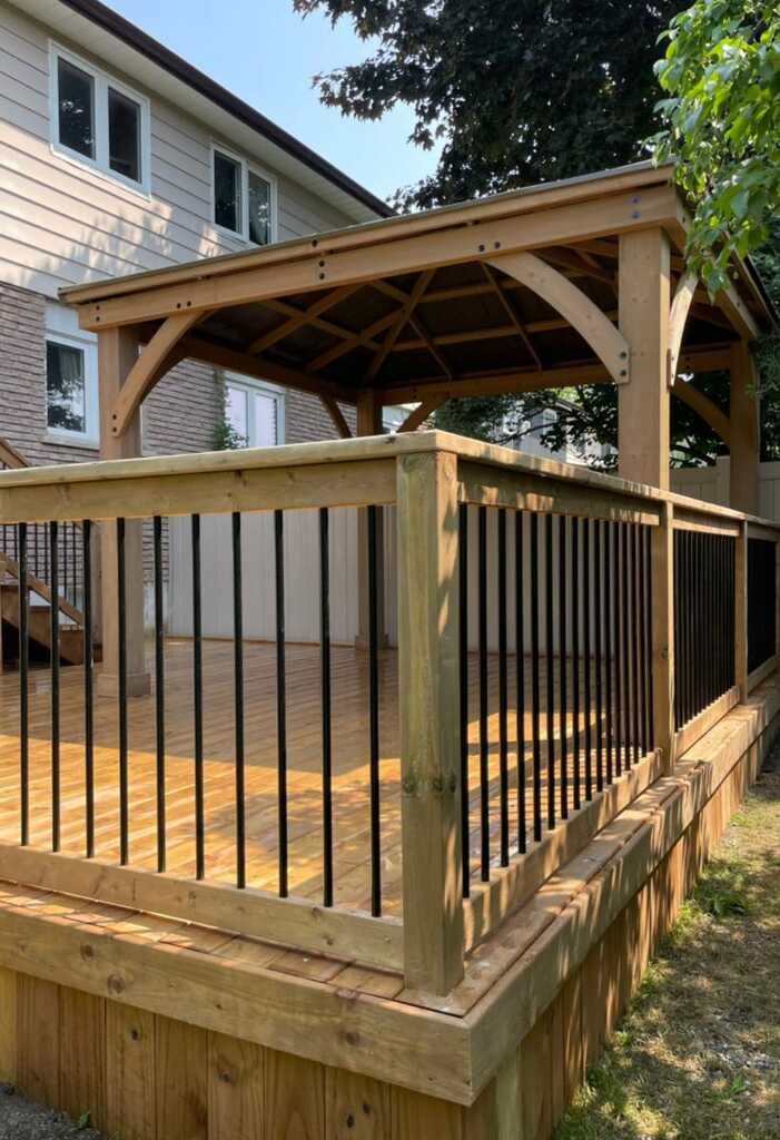 Wooden deck with gazebo. Black railing, brown gazebo, and tan house in the background.