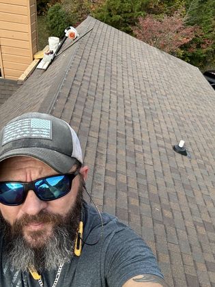 Man with beard and sunglasses on a shingled roof, holding a phone for a selfie.