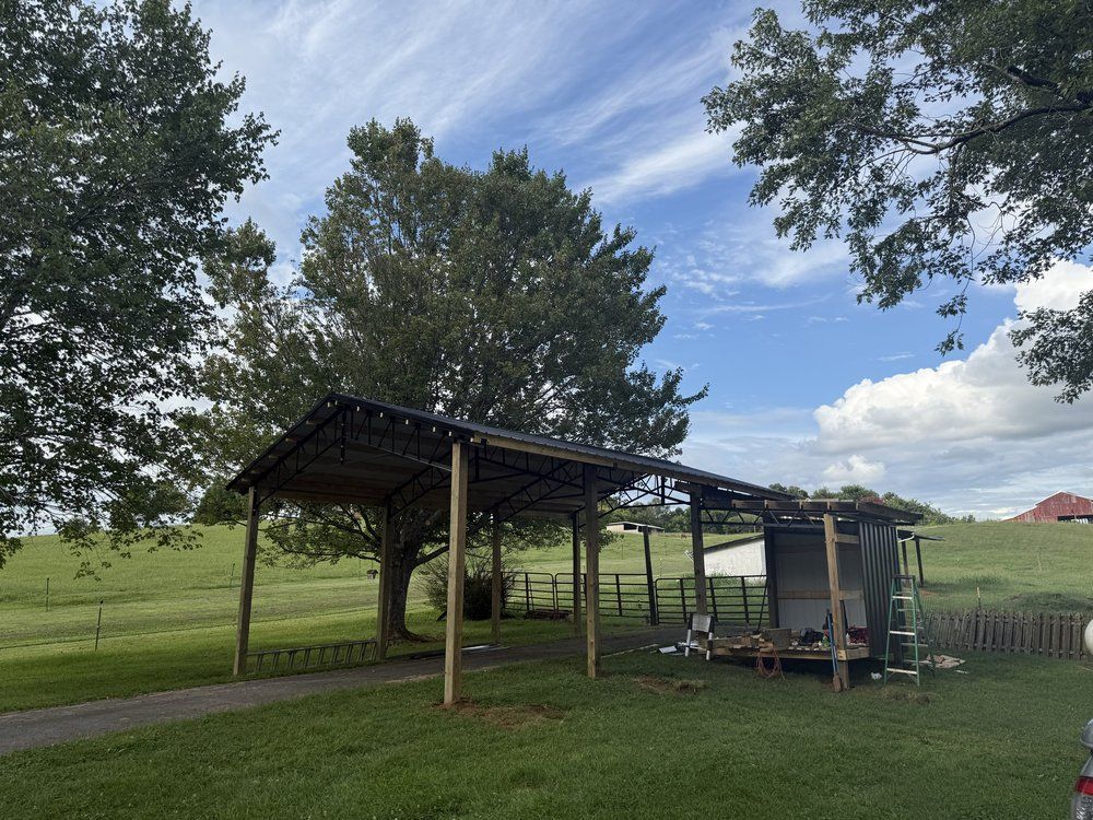 A rustic shed with a covered area, surrounded by green grass, trees, and a blue sky with clouds.